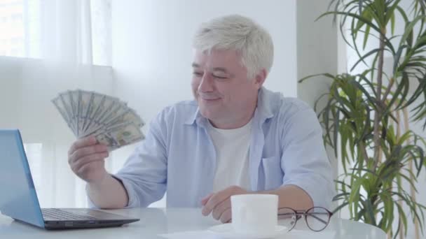 Feliz hombre mayor sentado en la mesa sosteniendo un paquete de dólares y sonriendo a la cámara. Retrato de un hombre de negocios caucásico satisfecho y maduro alardeando de riqueza. Freelancer masculino trabajando en línea. — Vídeos de Stock