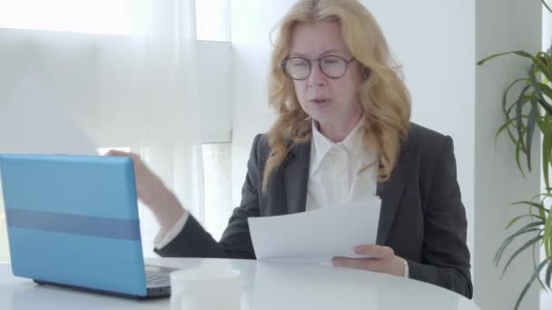 Serious Caucasian mid-adult businesswoman in eyeglasses talking on selfie camera on laptop as camera moves down to feet in funny unicorn slippers. Advantages of remote working on Covid-19 pandemic. — Stock Video