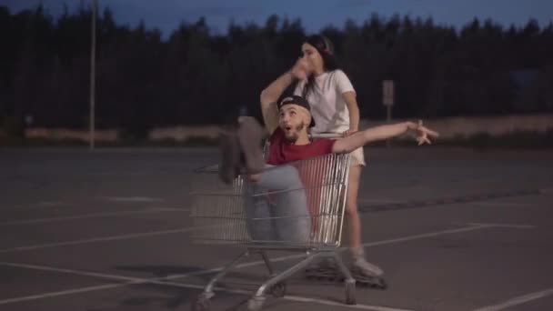 Fröhliche junge Frau auf Rollschuhen reitet fröhlichen Mann im Einkaufswagen. Porträt eines glücklichen kaukasischen Paares, das sich abends im Freien amüsiert. Lustige Teenager-Freizeit. — Stockvideo