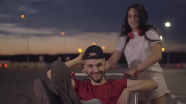 Portrait de beau jeune homme assis dans le panier et souriant belle femme tenant chariot. Joyeux petit ami caucasien et petite amie s'amuser sur le parking vide dans la soirée. — Video