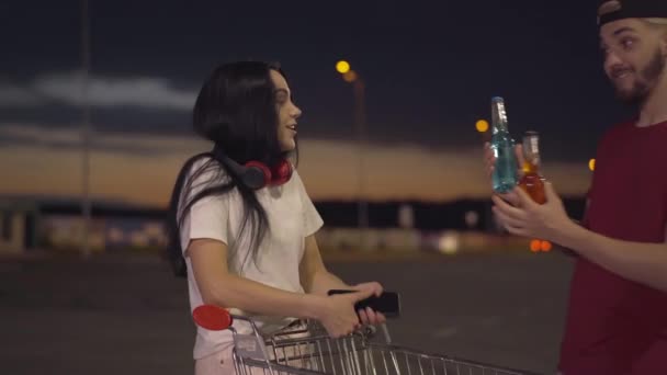 Middle shot of joyful young woman standing on empty parking lot with trolley at night as cheerful man coming with colorful beer. Portrait of relaxed carefree Caucasian couple resting in urban city.