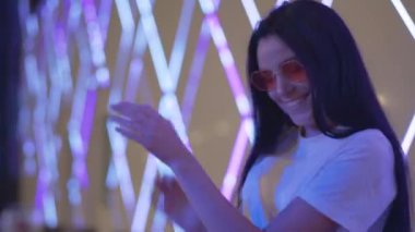 Güneş gözlüklü, neon ışıklarla dans eden mutlu, rahat beyaz kadın portresi. Neşeli gülümseyen genç dansçı gece kulübünde dinleniyor. Pozitif güzel kadın eğleniyor..