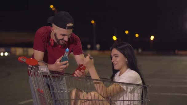 年轻迷人的女人坐在推车上，与英俊而快乐的男人叮咬着啤酒。无忧无虑的白人夫妇夜晚在空旷的城市停车场玩乐的肖像. — 图库视频影像