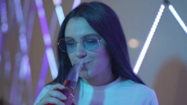 Gece kulübünde bira içen ve güneş gözlüğünü çıkaran tasasız genç bir kadın portresi. Güzel beyaz bir bayanın neon ışıklarda alkolle eğlendiği yakın plan çekimler. Milenyum nesli.