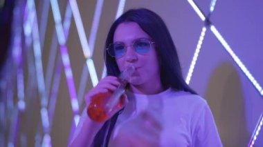 Genç kaygısız kadının renkli alkol içip gece kulübünde kameraya gülümsediği bir kare. Partide güneş gözlüklü, neon ışıklarıyla poz veren, pozitif milenyum bayanı portresi.