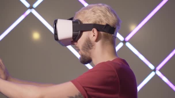 Vue latérale d'un jeune homme excité en lunettes VR touchant un mur invisible. Gros plan d'un joueur souriant et joyeux en réalité virtuelle dans VR-club. Millennial gars avoir du plaisir debout dans les néons. — Video