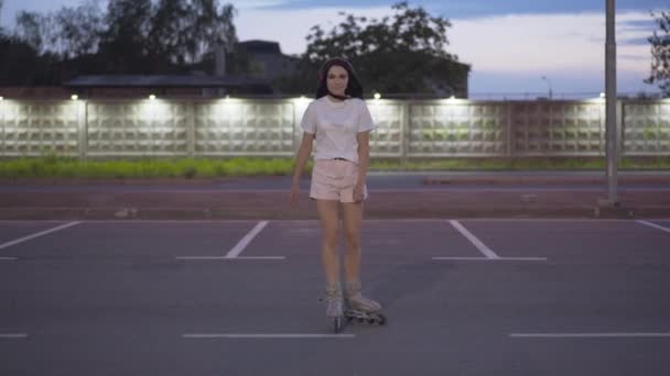 若い屈託のない女性のローラーでカメラに乗っている。夕暮れ時に楽しむ喜びの白人スケーターの肖像画。幸せな笑顔女性スケート上の空駐車場で街. — ストック動画