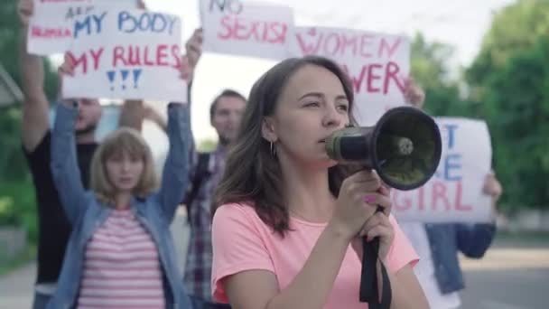 Retrato de una activista feminista gritando a través de un megáfono con gente agitando pancartas al fondo. Personas caucásicas conscientes que apoyan los derechos de la mujer y la igualdad de género. Huelga de feminismo. — Vídeo de stock