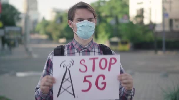 Portrett av en hvit mann med Stop 5G-banner i bygata. Alvorlig mannlig demonstrant i ansiktsmaske som protesterte mot mobile tårn på Covid-19 pandemien. Opptøyer. – stockvideo