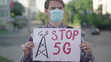 Kamera, Beyaz Maskeli Adam 'ın elindeki 5G plaketine yaklaşıyor. Mobil kulelerin radyoaktif kirliliğini protesto eden erkek eylemci Covid-19 solunum hastalığına yol açıyor.