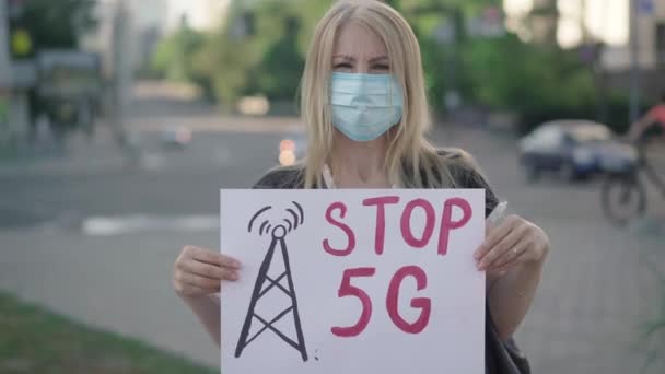 Yüz maskeli sarışın beyaz kadın 5G mobil kulesini protesto ediyor. Rack kadın aktivistten sloganı Stop 5G olan pankarta geçiyor. Radyasyona karşı bireysel isyan Covid-19. — Stok video