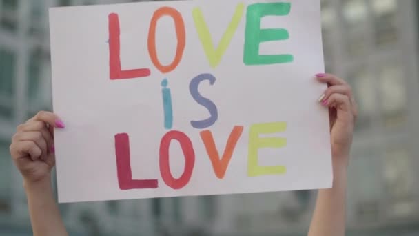 Close-up de amor levantado é bandeira do amor em mãos femininas caucasianas. Ativista lésbica irreconhecível segurando cartaz com slogan contra a homofobia. jovem mulher no gay orgulho desfile. — Vídeo de Stock