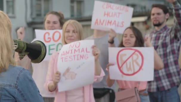 Vista posterior de la joven mujer caucásica gritando a través del megáfono con personas borrosas apoyando la manifestación contra la violación de los derechos de los animales en segundo plano. Líder de protesta alentando a eco-activistas. — Vídeo de stock