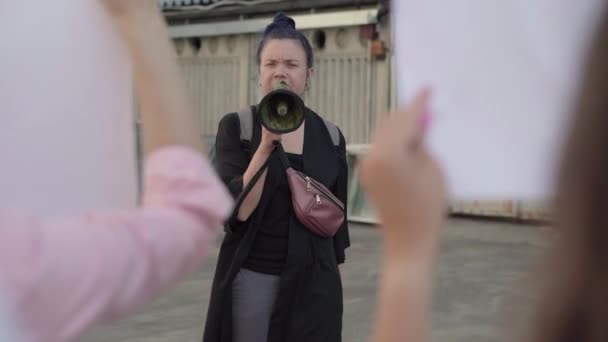 Retrato de líder de protesto gritando através do megafone como bandeiras agitando multidão irreconhecível. Discurso emocional da mulher caucasiana em manifestação. Ativista incentivando manifestantes na cidade urbana. — Vídeo de Stock