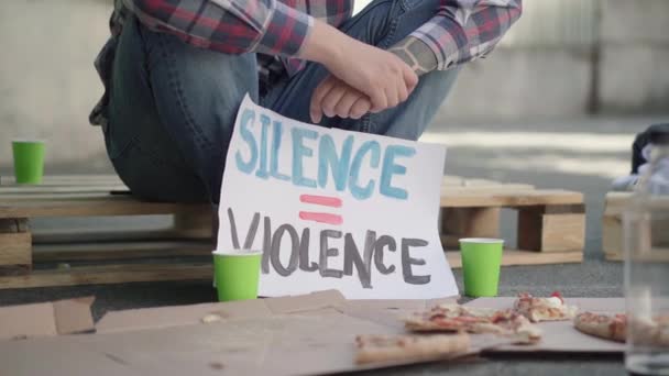 Närbild av Tystnad är våld banner med oigenkännlig manlig protestant sitter på pall i bakgrunden. Vit man med plakat mot kön eller etnisk ojämlikhet. — Stockvideo