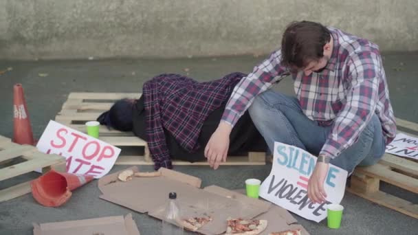 Utmattade aktivister sover på lastpallar efter demonstrationen. Porträtt av ung vit man och kvinna med antirasism eller jämställdhet banners utomhus med halväten pizza. — Stockvideo