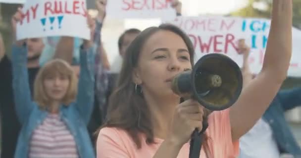 Jonge mooie protestleider schreeuwend door megafoon en gebaren makend met mensen die feministische posters op de achtergrond schudden. Kaukasische demonstranten die gendergelijkheid verdedigen. Cinema 4k ProRes Hoofdkwartier. — Stockvideo