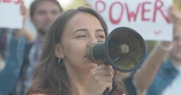 Close-up da jovem feminista caucasiana gritando através do megafone. Líder do protesto encorajando manifestantes com cartazes anti-sexismo em segundo plano. Pessoas que protestam contra a desigualdade. Cinema 4k ProRes HQ. — Vídeo de Stock
