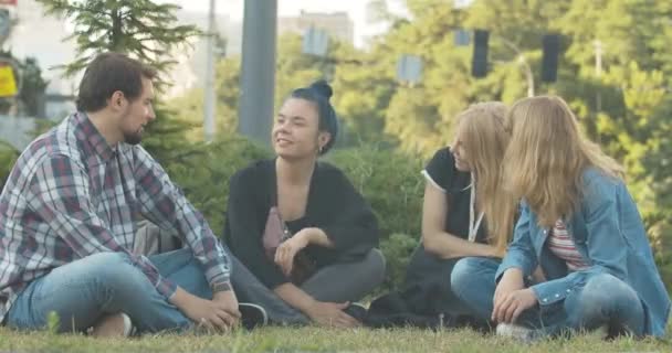 Négy ember ül a zöld füvön a városi parkban és beszél. Portré pozitív kaukázusi férfiakról és nőkről, akik a szabadban beszélgetnek a napsütéses nyári napon. Szabadidő-koncepció. Cinema 4k ProRes főhadiszállás. — Stock videók