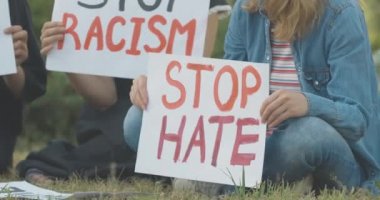 Kadın ellerindeki nefret pankartından vazgeç. Çimlerin üzerinde ırkçılık karşıtı afişlerle oturan tanınmamış genç beyaz protestocu. Sosyal eşitsizliği protesto eden bir kadın. Sinema 4k ProRes Merkezi.
