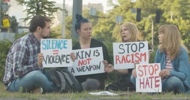 Cuatro manifestantes caucásicos sentados en el césped con pancartas antirracistas y hablando. Grupo de hombres y mujeres caucásicos protestando contra la desigualdad social al aire libre. Protesta de brazos caídos. Sede del cine 4k ProRes. — Vídeos de Stock