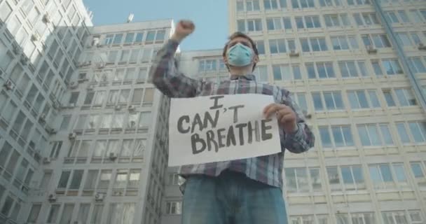 Vista inferior del manifestante entusiasta con máscara facial sosteniendo No puedo respirar pancarta y gesto. Joven hombre caucásico protestando contra la discriminación racial en la pandemia de Covid-19. Sede del cine 4k ProRes. — Vídeo de stock