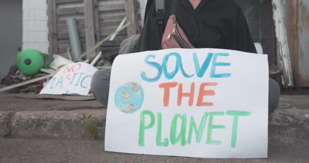 Felismerhetetlen női öko-aktivista ül a Mentsd meg a bolygót felirattal a háttérben szeméttel. Fehér nő tiltakozik a környezetszennyezés ellen. Cinema 4k ProRes főhadiszállás. — Stock videók