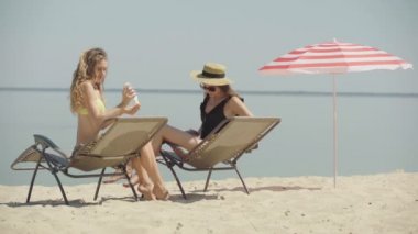 Kumlu sahilde bronzlaşmış cesede güneş kremi süren zayıf, pozitif genç kadınlar. Güneşli yaz tatilinde güneş kremi süren kendine güvenen beyaz turistlerin portresi. Cilt bakımı kavramı.