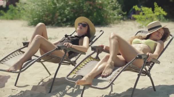 Atrakcyjna młoda kobieta budzi się jako przyjaciółka śpiąca na leżaku na piaszczystej plaży. Portret dwóch pozytywnych białoruskich pięknych turystów odpoczywających na letniskowym kurorcie na wakacjach. Turystyka i styl życia. — Wideo stockowe