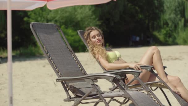 Όμορφη νεαρή Καυκάσια γυναίκα απολαμβάνει τις διακοπές στην αμμώδη παραλία ως φίλος έρχεται και κάθεται κάτω στην ξαπλώστρα. Πορτραίτο δύο λεπτών τουριστών που αναπαύονται στο θερινό θέρετρο. Έννοια τουρισμού. — Αρχείο Βίντεο
