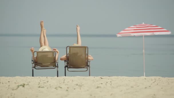 Duas jovens mulheres irreconhecíveis tremendo bronzeado pernas finas na praia de areia. Ampla vista panorâmica de turistas caucasianos descansando em espreguiçadeiras no resort de verão. Senhoras felizes desfrutando de férias. — Vídeo de Stock