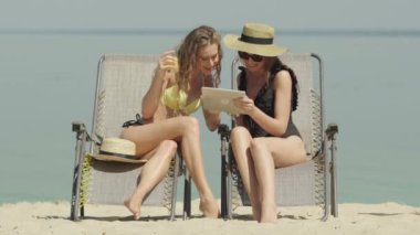 Tabletle kumsalda konuşan mayolu iki mutlu genç kadının portresi. Güzel, kendine güvenen beyaz turistler yaz tatilinin tadını çıkarıyorlar. Zengin kadınlar güneşlenirken güneşleniyor..