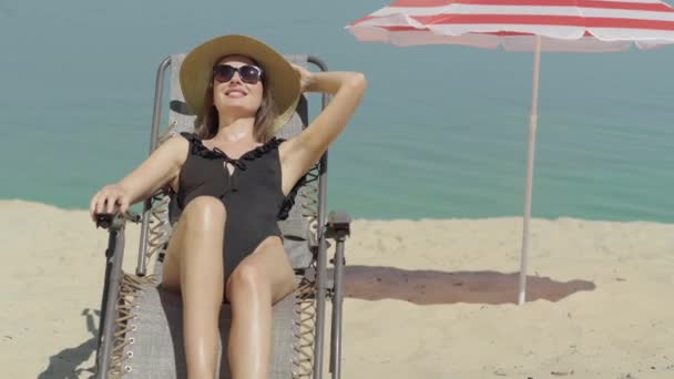 Joyeux jeune femme en maillot de bain et lunettes de soleil reposant sur une chaise longue sur une plage de sable. Portrait d'un magnifique bain de soleil touristique souriant dans une station balnéaire ensoleillée. Concept de tourisme. — Video