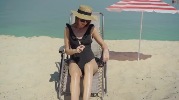 Caméra approche sensuelle jeune femme caucasienne pulvérisation écran solaire à la station estivale ensoleillée. Portrait de magnifique bronzage touristique confiant sur une chaise longue sur une plage de sable. — Video