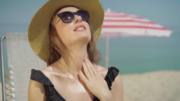 Retrato de mujer relajada feliz frotando protector solar en el pecho. Joven hermosa dama caucásica aplicando protector solar en la playa de arena y sonriendo. Seguro delgado turista tomar el sol en el soleado complejo de verano. — Vídeos de Stock