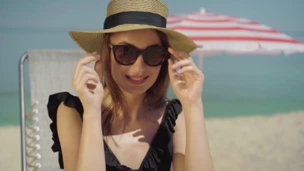 Portrait de femme caucasienne magnifique positive dans les lunettes de soleil clin d'oeil à la caméra et souriant. Heureux jeune beau touriste profitant de vacances sur la plage de sable fin à la station balnéaire ensoleillée. — Video