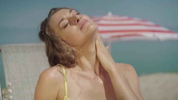 Jeune femme séduisante appliquant de la crème solaire sur le cou et les épaules. Portrait de sensuel bain de soleil touristique caucasien sur la plage de sable fin par une journée ensoleillée. Magnifique dame au repos à la station estivale. — Video