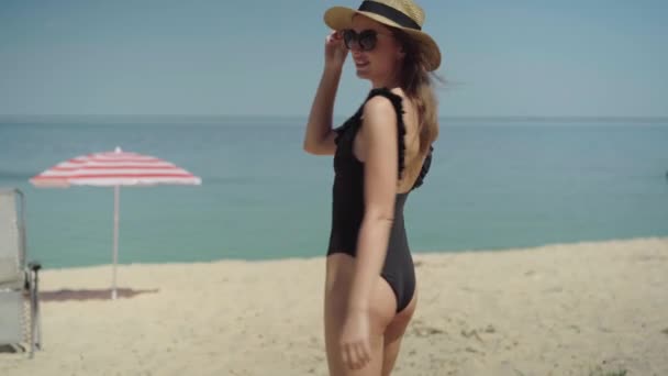 相机跟随着自信的黑发女人，戴着太阳镜，头戴帽子，走向大海。年轻瘦弱的游客漫步在沙滩上,面带微笑.积极快乐的女士在避暑胜地休息. — 图库视频影像