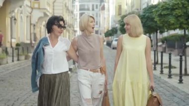 Olumlu orta yaşlı kadınlar modern Avrupa şehrinde kaldırımlarda gezinip konuşuyorlar. Güneşli bir yaz gününde dışarıda yürüyüp sohbet eden neşeli beyaz arkadaşlarının portresi..