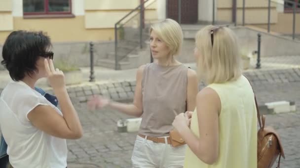 Vékony, középkorú nő beszélget a barátaival az európai városi utcában. Portré három kaukázusi nőről, akik a szabadban pletykálnak a napsütéses nyári napon. Barátság és szabadidő koncepció. — Stock videók
