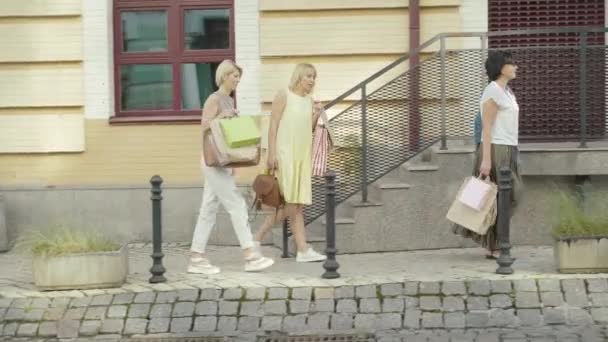 Vista lateral de compradores caucásicos de mediana edad que pasean por la calle de la ciudad el viernes negro. Amplio tiro de mujeres felices con bolsas de compras caminando al aire libre en el día soleado y charlando. — Vídeos de Stock