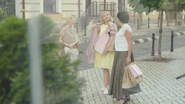 Amici ammirando nuova sciarpa di positivo bella donna di mezza età sulla strada della città. Donne caucasiche di mezza età che discutono di acquisti all'aperto con borse della spesa. Cespuglio verde che passa davanti. — Video Stock