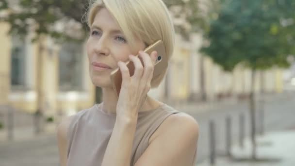 Primer plano de la mujer caucásica de mediana edad que camina por la calle de la ciudad y habla por teléfono. Retrato de la señora rubia elegante feliz disfrutando de un soleado día de verano al aire libre. — Vídeo de stock