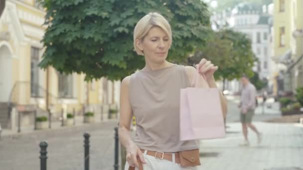 Retrato del feliz comprador medio adulto estirando la bolsa de compras a la cámara y sonriendo. Mujer rubia caucásica segura de mediana edad parada en la calle urbana de la ciudad en el soleado día de verano el viernes negro. — Vídeos de Stock