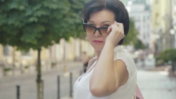 Retrato de cerca de una mujer caucásica adulta confiada que se pone gafas de sol y sonríe a la cámara. Mujer hermosa posando con bolsas de compras en la soleada calle de la ciudad de verano. — Vídeos de Stock