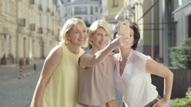 Mutlu sarışın, orta yaşlı kadın arkadaşlarıyla gün ışığında selfie çekiyor. Yazın açık havada dinlenen üç beyaz kadının portresi. Kentsel şehirde kendine güvenen modern kadınlar.