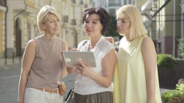 Ritratto di tre allegre donne di mezza età in piedi alla luce del sole sulla strada della città e utilizzando tablet. Buoni turisti caucasici che scelgono posto per visitare la città straniera. Concetto turistico estivo. — Video Stock