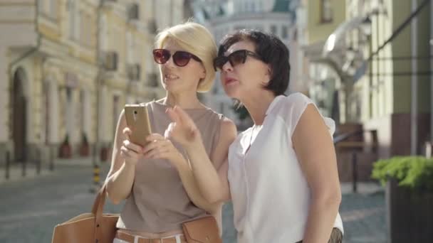 Donne bionde e brune di mezza età che scattano foto sulla strada della città straniera nella soleggiata giornata estiva. Ritratto di due turisti caucasici positivi che riposano in Europa. Turismo e viaggi. — Video Stock