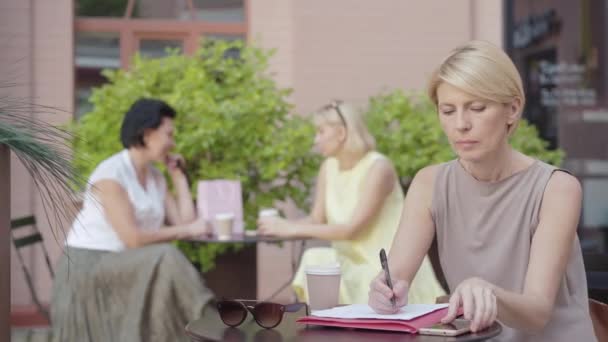 Retrato de una mujer de negocios seria y confiada sentada en una cafetería al aire libre con documentos y teléfonos inteligentes y mujeres borrosas descansando en el fondo. Hermosa mujer de negocios rubia trabajando en exceso en el descanso. — Vídeos de Stock