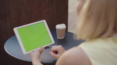 Kromakey ekranlı tablet tutan ve kahve içen tanınmamış bir kadın. Sarışın, beyaz, yetişkin bir kadın internette bir şey izliyor. Kablosuz iletişim, internette sörf..